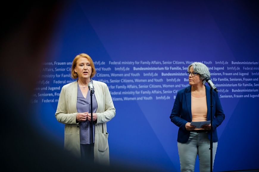 Lisa Paus und Kerstin Claus stehen vor einer blauen Wand, vor ihnen sind Mikrofone aufgestellt